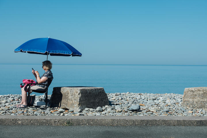 A woman sits under an umbrella looking at her phone beside the sea